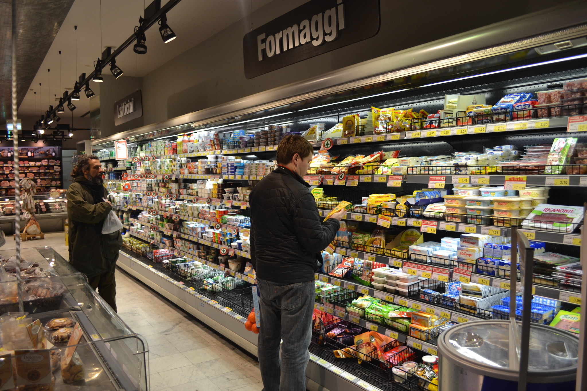 Un hombre se para frente a un gabinete enfriador en un supermercado.  Sobre su cabeza, el letrero dice Formaggi